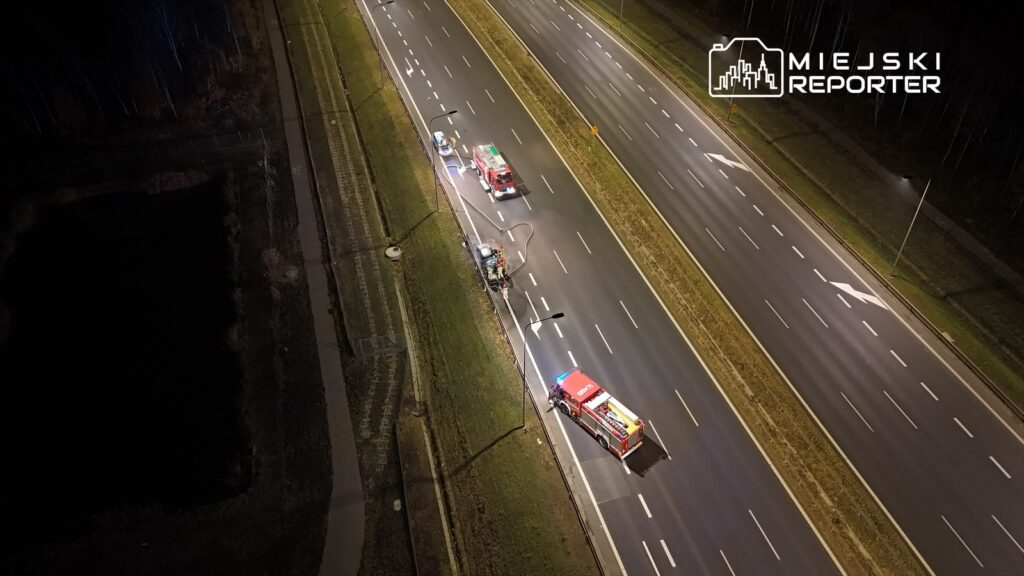 Strażacy przy akcji ratunkowej na poboczu drogi, obok zaparkowanego wozu strażackiego w nocy.