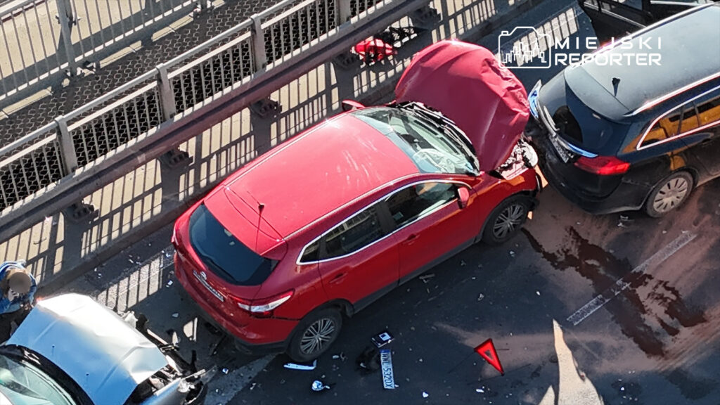 Czerwony samochód osobowy z uszkodzoną maską stoi na jezdni, obok niego leży rozbite szkło i plama płynów.
