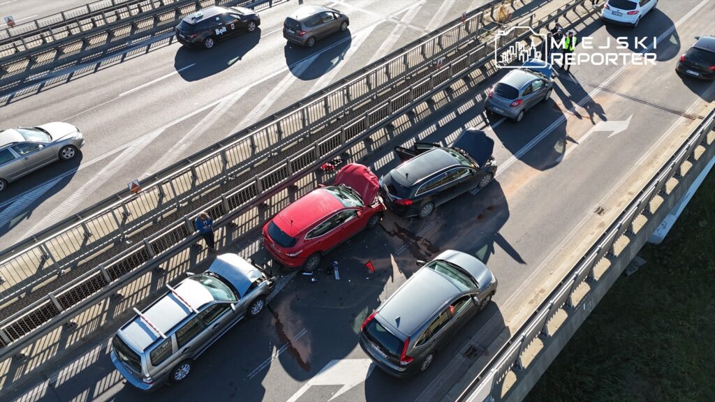 Wielokrotny wypadek drogowy z udziałem kilku samochodów na ruchliwej jezdni, z uszkodzonymi pojazdami i oznaczeniem ostrzegawczym.