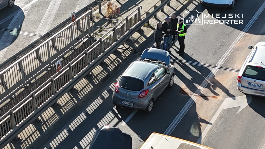 Kierowca w szarym samochodzie rozmawia z policjantami na ruchliwej drodze, obok otwartej maski pojazdu.