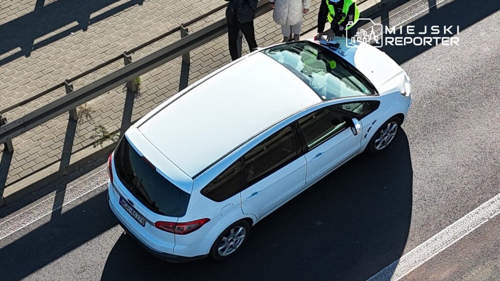 Policjant w kamizelce odblaskowej sprawdza dokumenty kierowcy w białym samochodzie na ulicy.