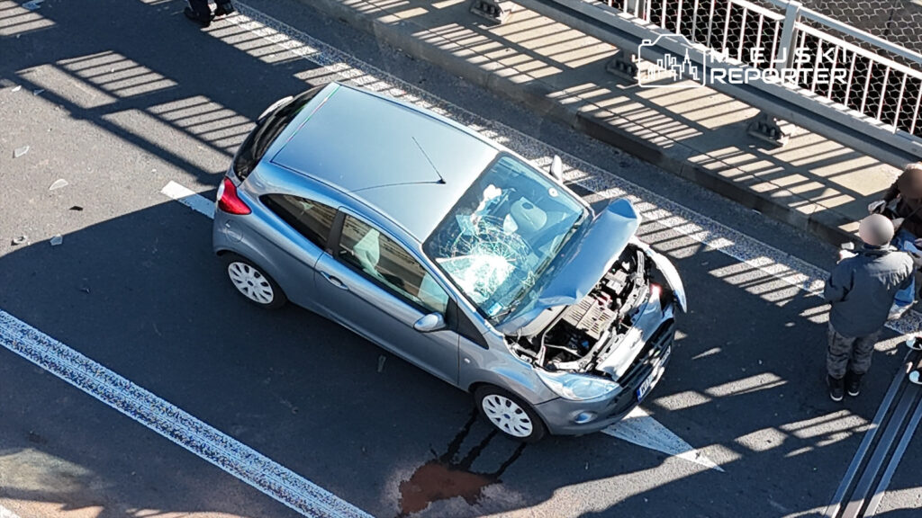 Srebrny samochód osobowy z rozbitą szybą i otwartą maską stoi na jezdni, obok dwie osoby rozmawiają.