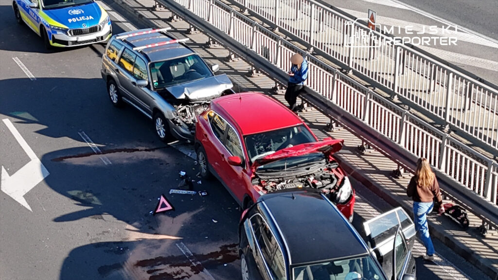 Zderzenie dwóch samochodów osobowych na jezdni, obok stoi policja oraz osoby zajmujące się sytuacją.