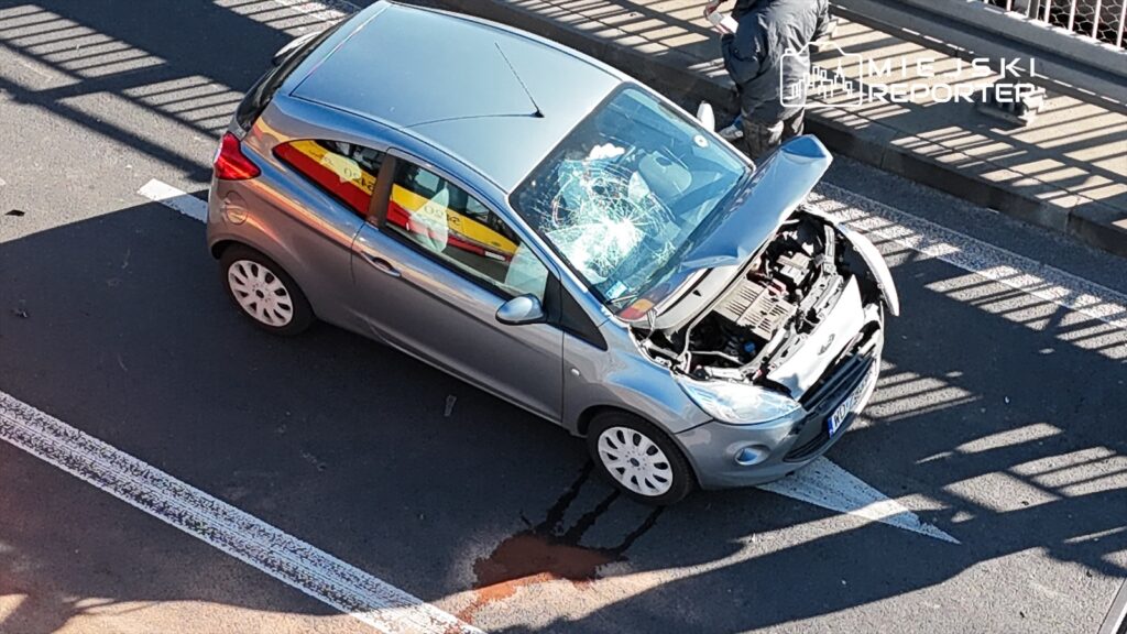 Samochód osobowy w kolorze srebrnym z uszkodzoną szybą i otwartą maską stoi na jezdni, obok widać plamę oleju.