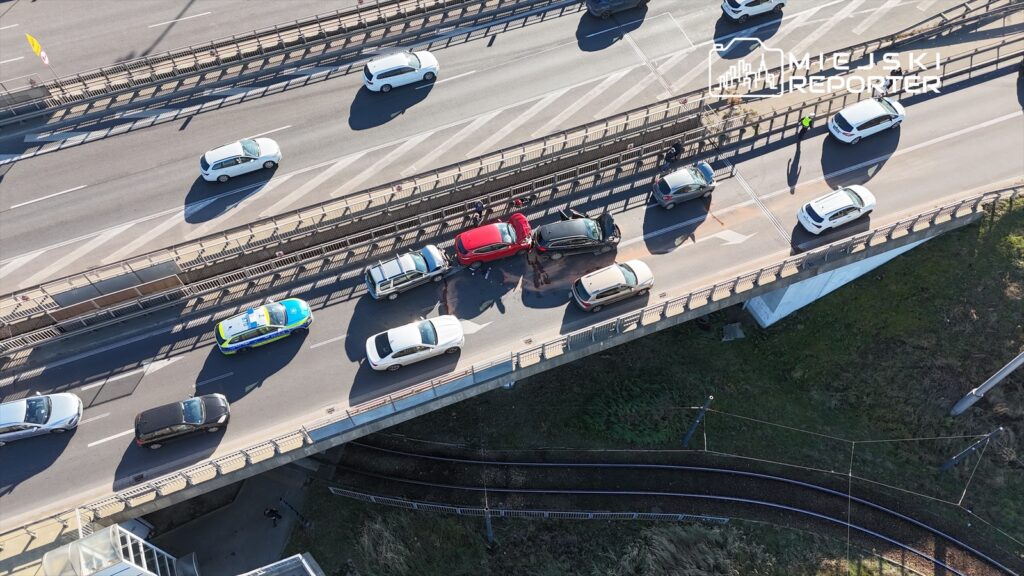 Cztery samochody zderzone na ruchliwej drodze, obok policyjny radiowóz oraz kilka osób pomagających poszkodowanym.