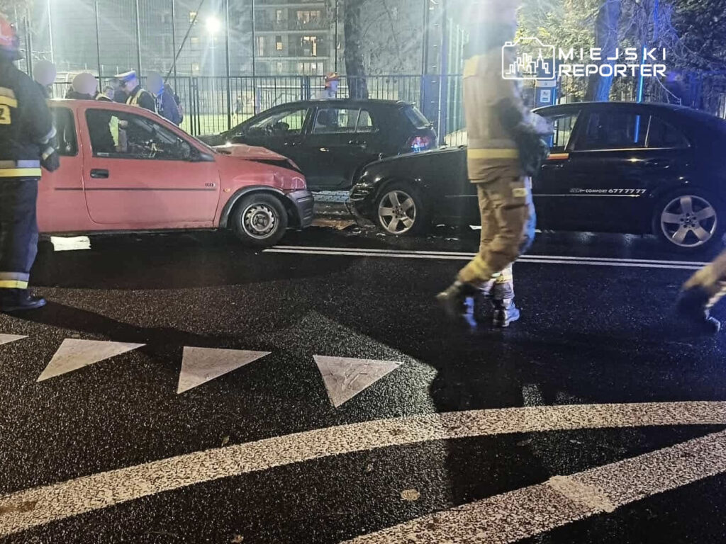 Grupa strażaków w mundurach przeprowadza działania na miejscu kolizji drogowej, w tle widoczne uszkodzone pojazdy.