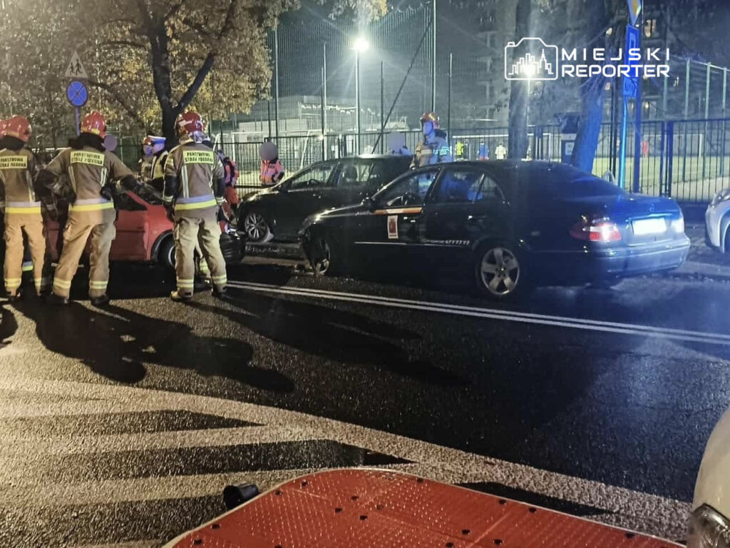 Grupa strażaków w mundurach, z hełmami, ocenia szkody po wypadku drogowym na oświetlonej ulicy, obok zaparkowanych samochodów.