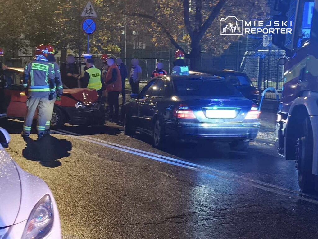 Na ulicy, zniszczony czerwony samochód stoi po zderzeniu z czarnym pojazdem, wokół zbierają się strażacy i policjanci.