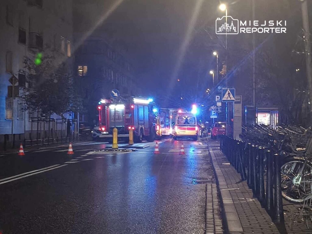 Na ulicy z widocznymi pojazdami straży pożarnej i ambulansami w nocy, z lampami błyskowymi i znakami ostrzegawczymi.