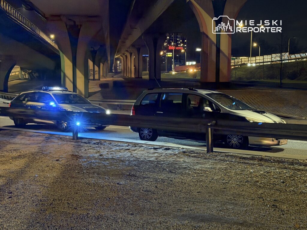 Policyjny radiowóz z włączonymi sygnałami świetlnymi zatrzymuje samochód osobowy pod estakadą w nocy.