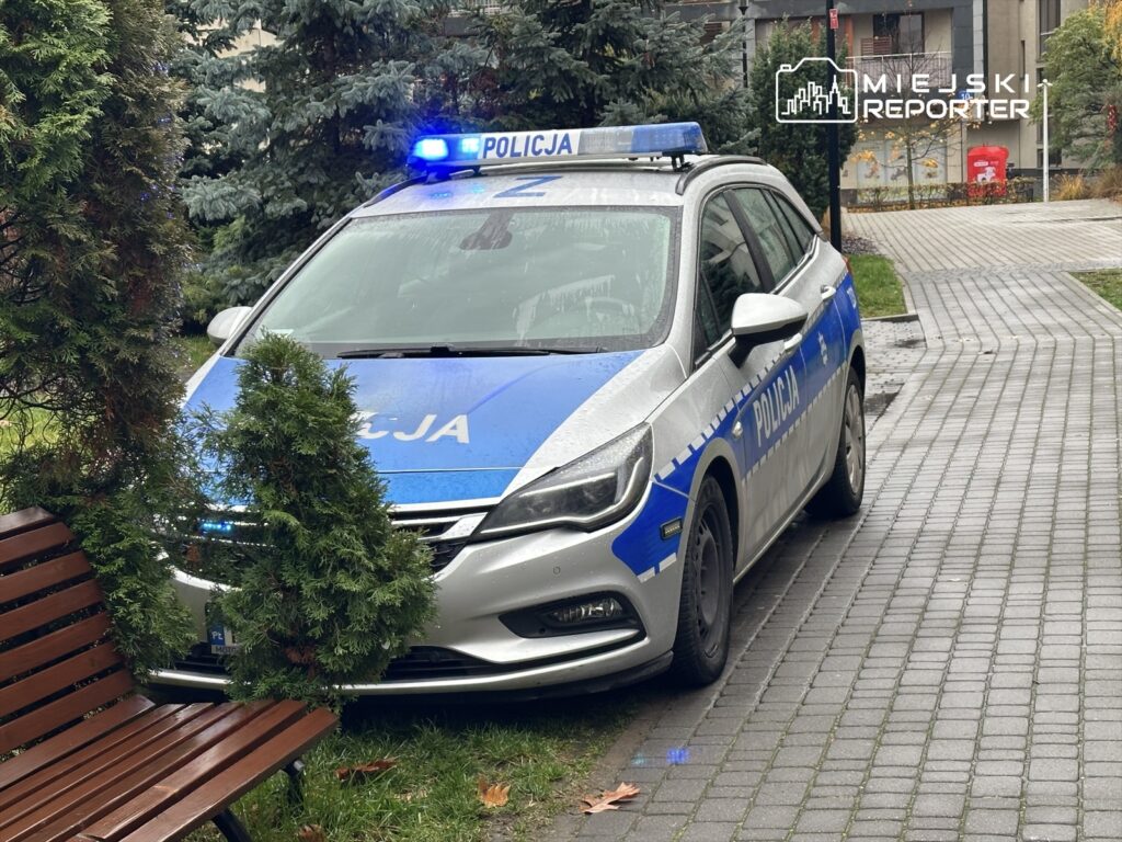 Policyjny radiowóz z włączonymi sygnałami świetlnymi zaparkowany na ścieżce w otoczeniu zieleni i drewnianej ławki.
