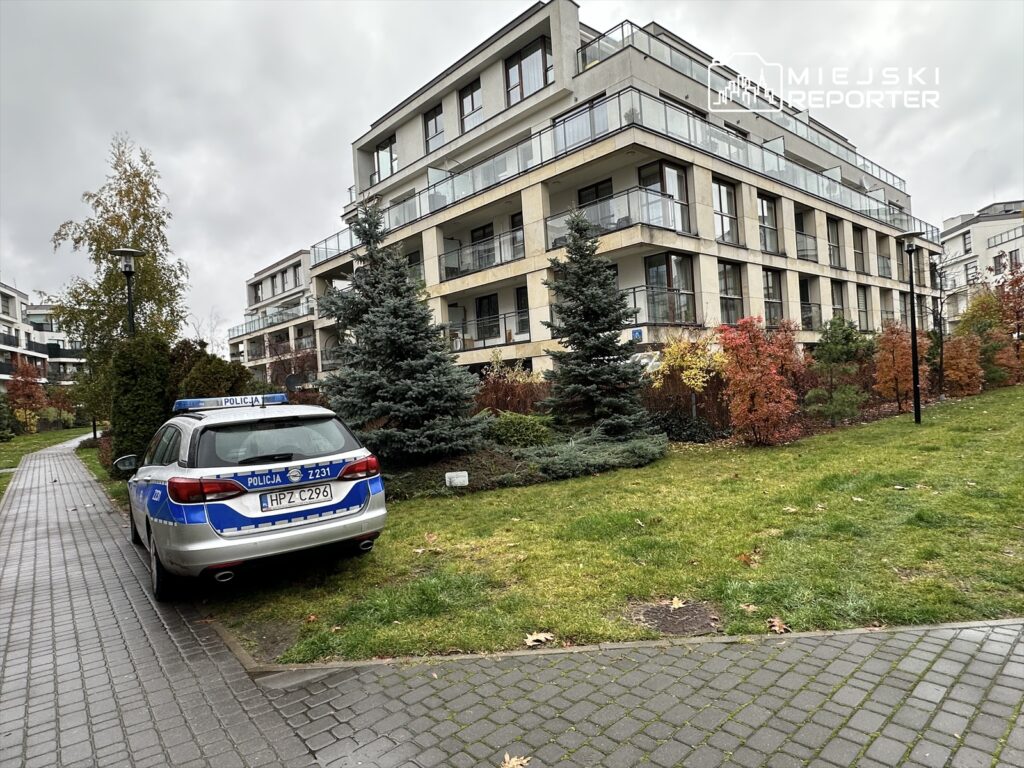 Policyjny samochód zaparkowany na trawniku przed nowoczesnym budynkiem mieszkalnym, otoczony zielenią.