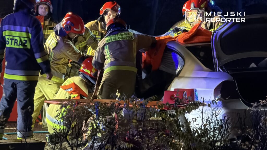 Grupa strażaków w specjalistycznym ubraniu prowadzi akcję ratunkową przy uszkodzonym samochodzie w nocy.