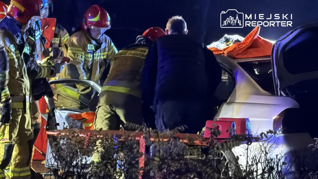 Grupa strażaków w mundurach i hełmach pracuje nad wydobyciem osoby z rozbitego samochodu w nocy.
