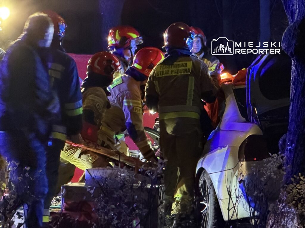 Grupa strażaków w hełmach udziela pomocy poszkodowanemu przy uszkodzonym samochodzie w nocy.