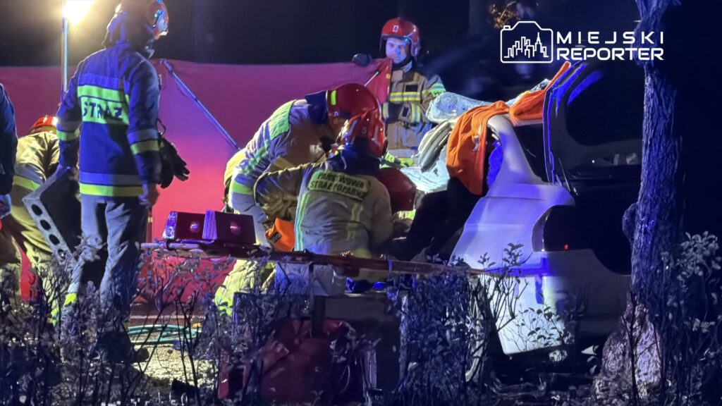 Grupa strażaków w mundurach ratuje osobę z rozbitego samochodu w ciemnym lesie, otoczona przez gałęzie i sprzęt ratunkowy.