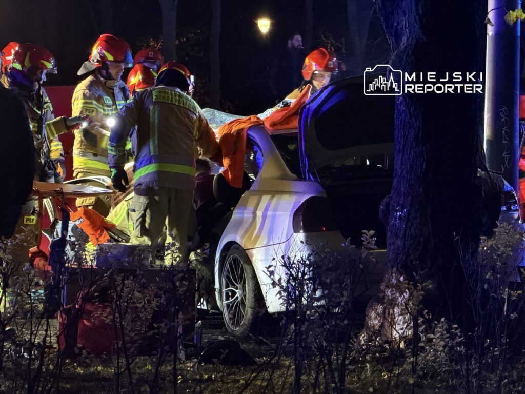 Grupa strażaków w kaskach pracuje nad usunięciem poszkodowanego z wraku samochodu w ciemnym otoczeniu.