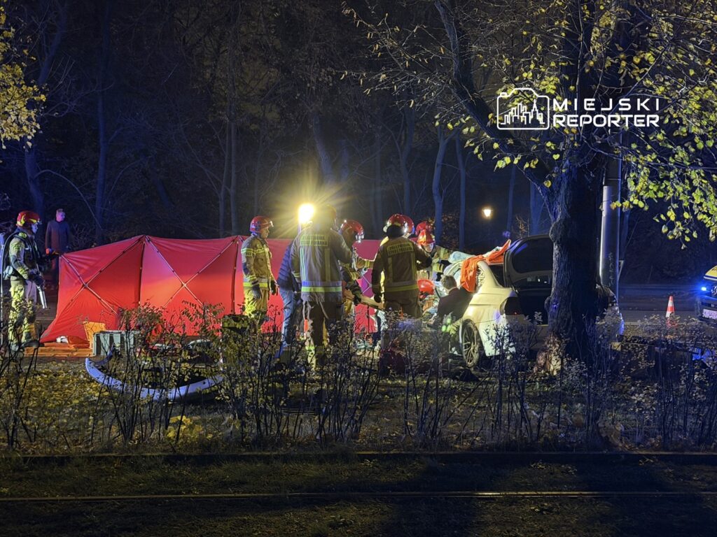 Grupa strażaków w kaskach pracuje przy wypadku drogowym, obok czerwonego namiotu oświetlonego przez latarki.