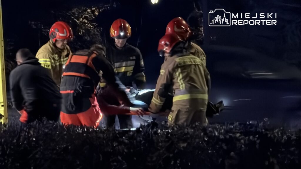 Ratownicy medyczni i strażacy udzielają pomocy poszkodowanemu na noszach w nocy, w miejskim otoczeniu.