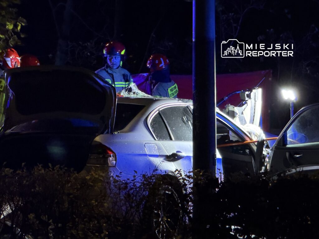Grupa strażaków w hełmach pracuje przy uszkodzonym samochodzie w nocy, oświetlonym przez lampy ratunkowe.