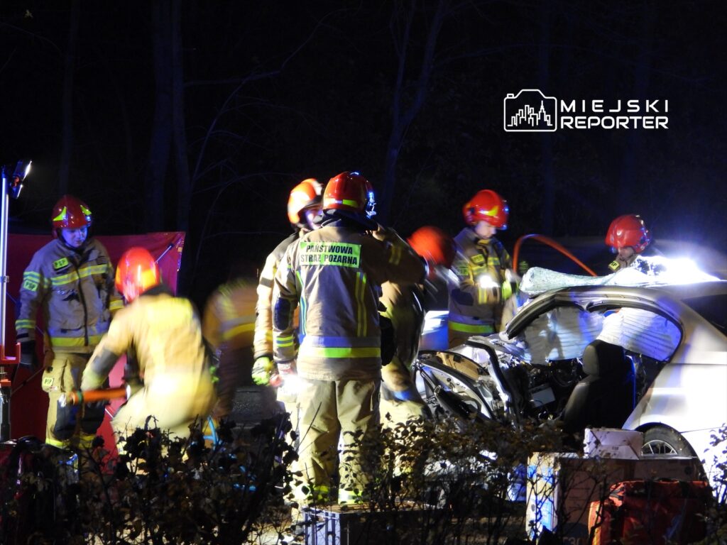 Grupa strażaków w hełmach i mundurach pracuje przy rozbitym samochodzie w ciemnym lesie, używając narzędzi ratunkowych.