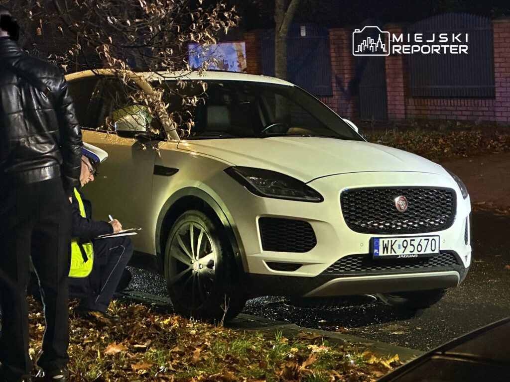 Funkcjonariusz policji notuje informacje obok białego samochodu marki Jaguar zaparkowanego na ulicy w nocy.