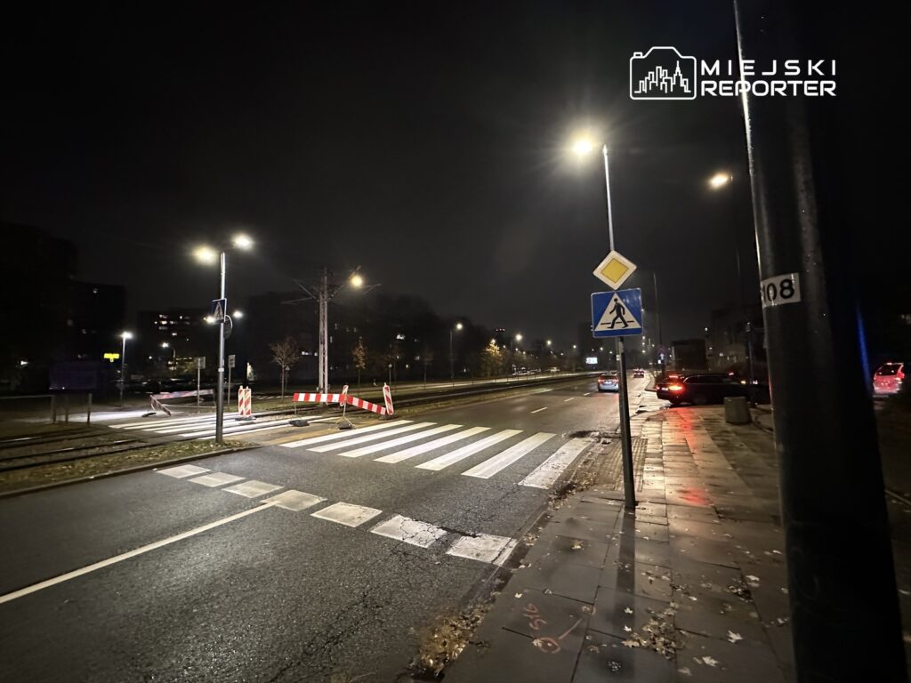 Oświetlona ulica nocą z przejściem dla pieszych, barierkami drogowymi i zaparkowanymi samochodami.