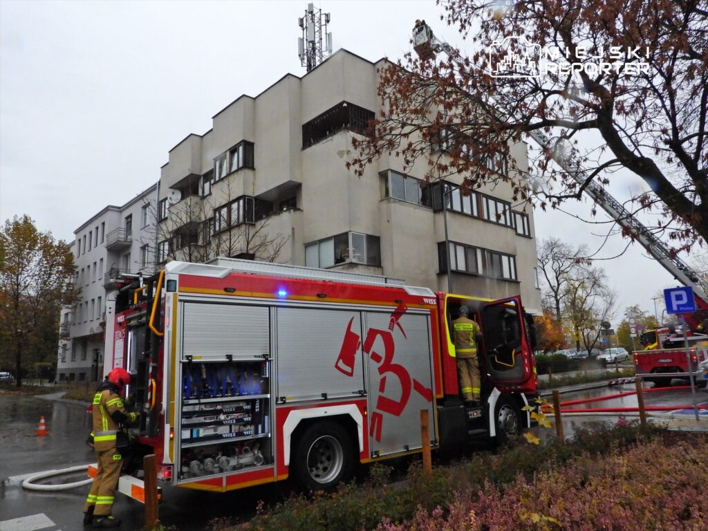 Strażacy obsługują wóz strażacki na tle budynku mieszkalnego w deszczową pogodę, z widocznymi drabinami.