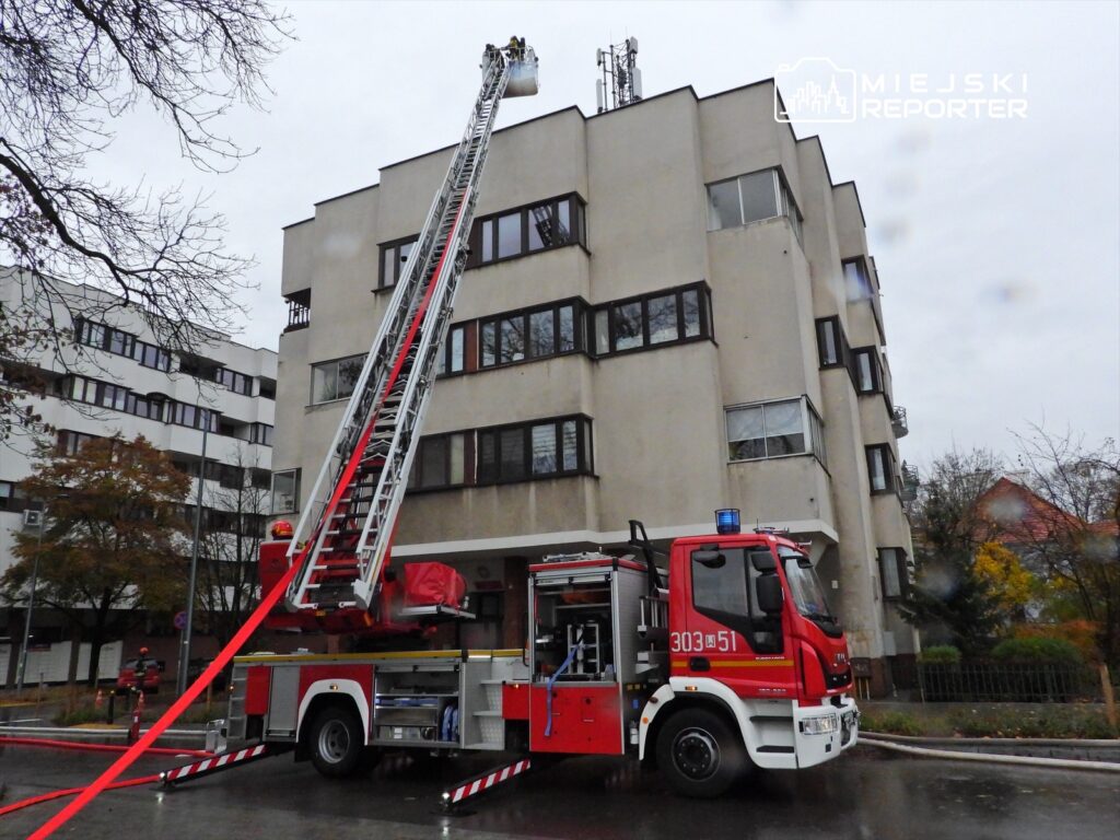 Strażacy na podnośniku w akcji ratunkowej przy budynku mieszkalnym, obok wóz strażacki z rozwiniętym wężem.