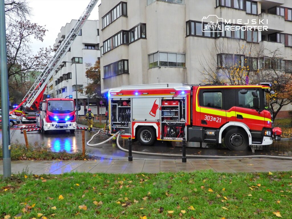 Strażacy przy wozie strażackim rozkładają sprzęt w pobliżu budynku mieszkalnego, obok podnośnika strażackiego.