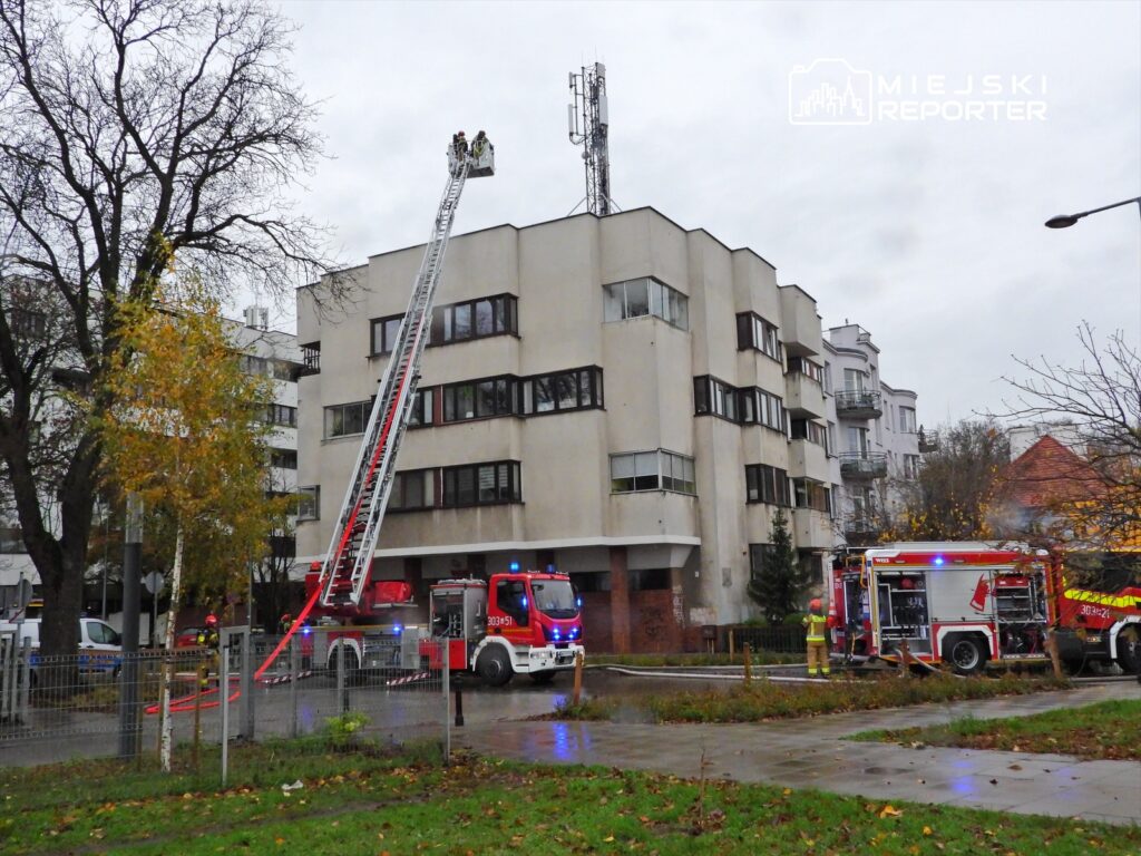 Strażacy na podnośniku gaszą pożar w oknie budynku mieszkalnego, obok parkują wozy strażackie.
