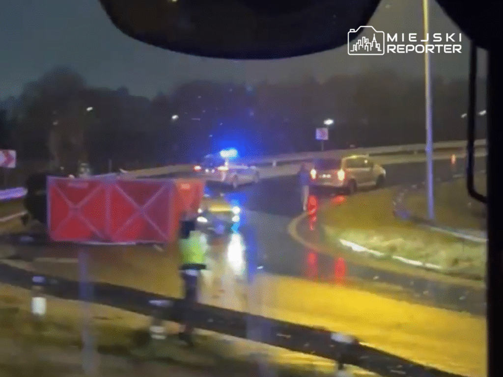 Na drodze widoczny jest przewrócony pojazd oraz radiowóz policyjny z włączonymi sygnałami świetlnymi.