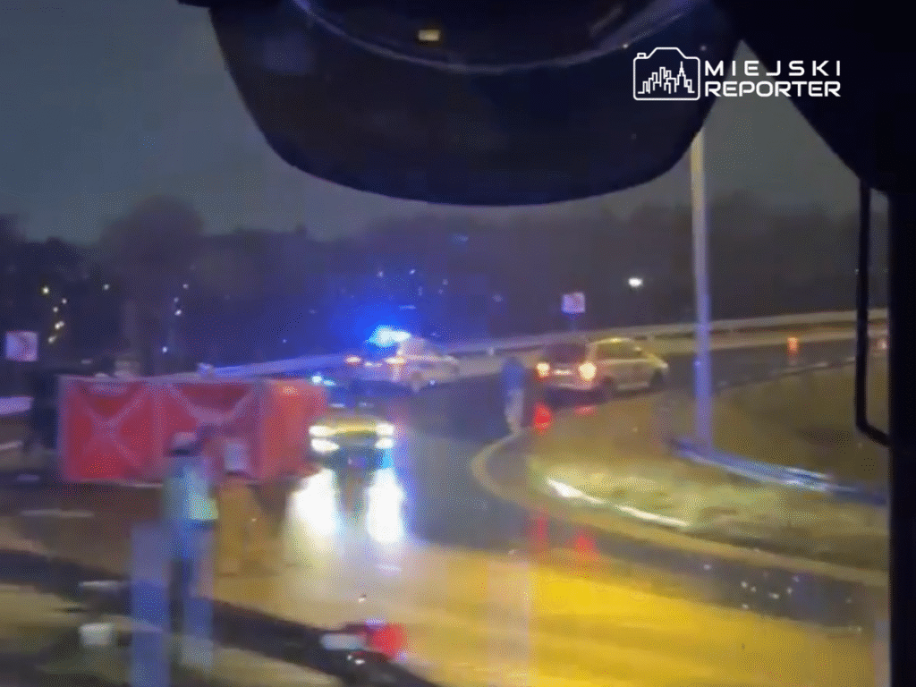 Na mokrej nawierzchni drogi stoją pojazdy służb ratunkowych, a jeden z samochodów jest przewrócony na bok.