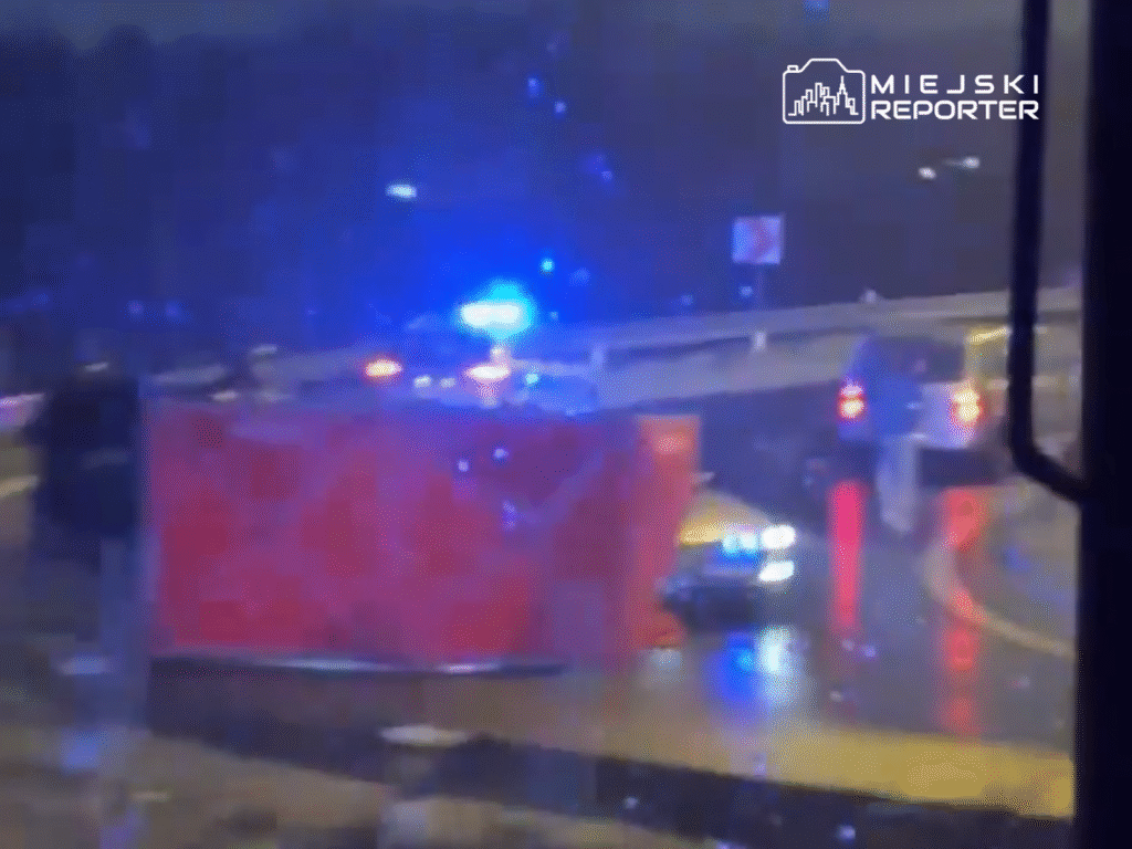 Policyjne samochody z włączonymi sygnałami świetlnymi zatrzymują ruch na mokrej drodze w deszczową noc.