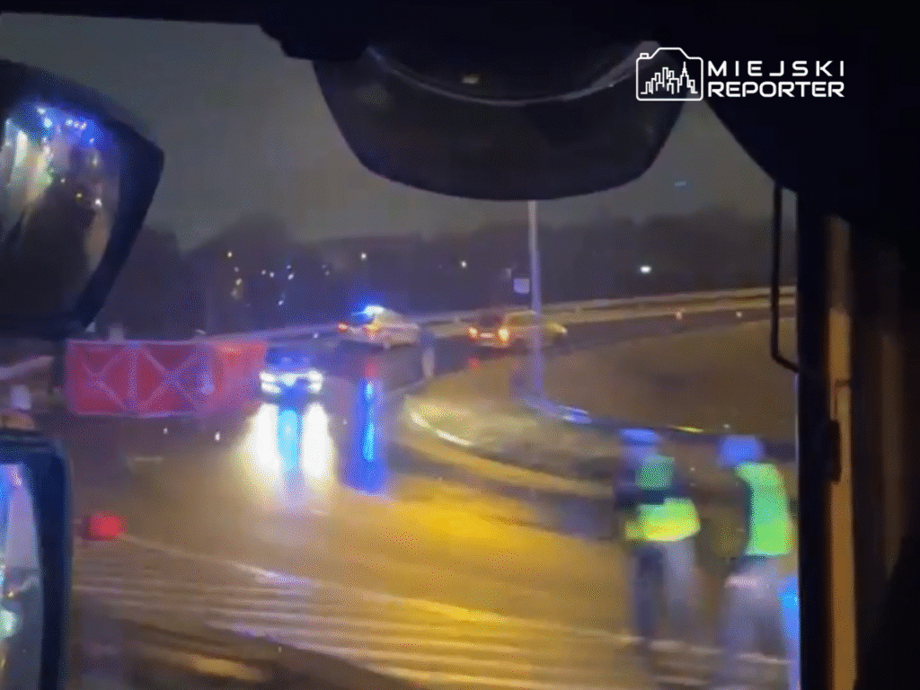 Policyjny radiowóz z włączonymi sygnalizatorami blokuje ruch na mokrej drodze, a obok pracujący mężczyźni w kamizelkach odblaskowych.