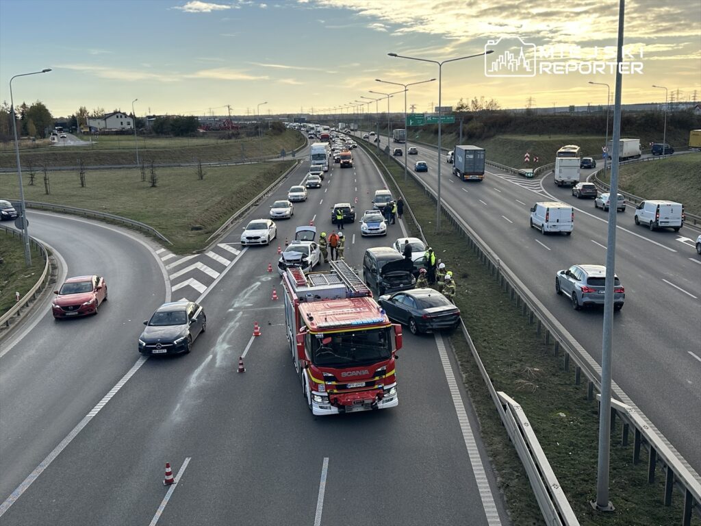 Na drodze nastąpił wypadek, w którym biorą udział dwa samochody osobowe; obok pracują strażacy i policja.