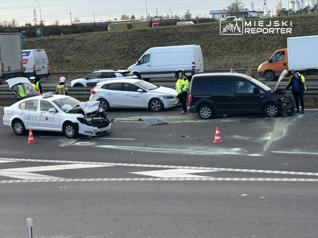 Na drodze stoją dwa uszkodzone samochody, w tym taksówka z wgniecionym przodem; obok pracują policjanci.