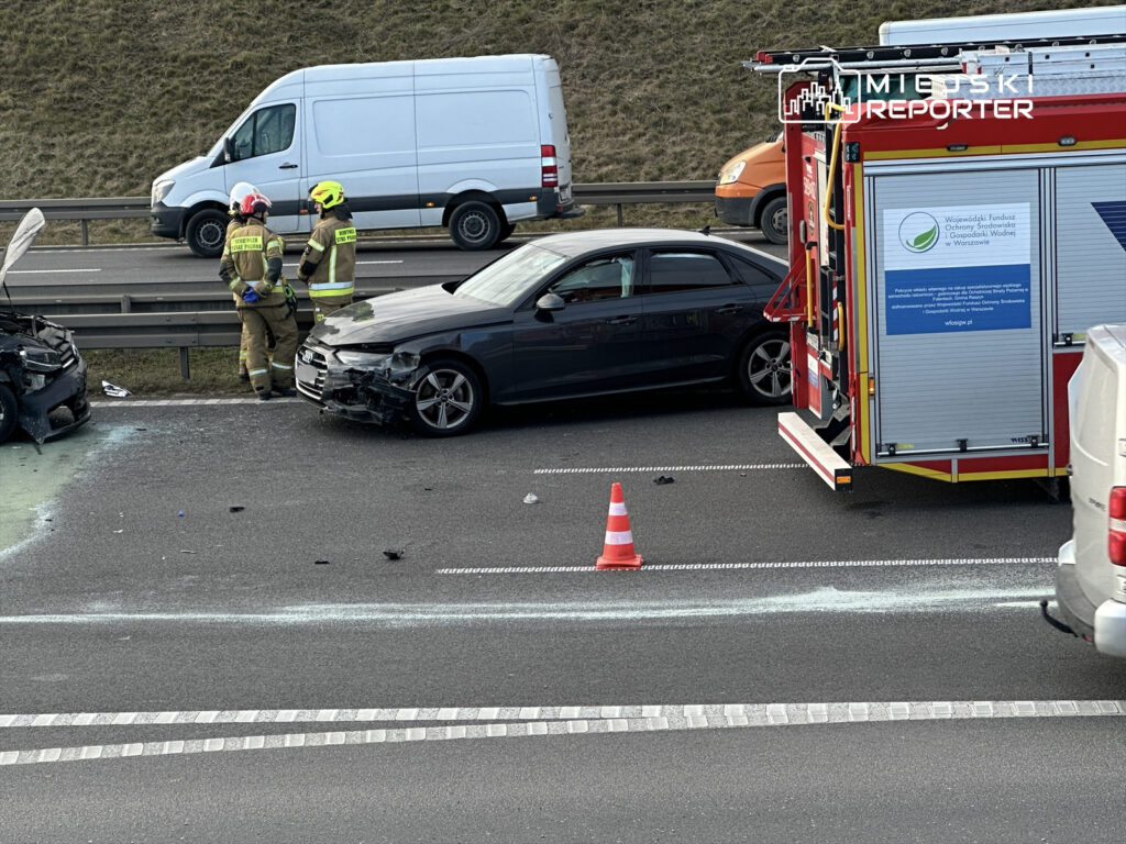 Dwóch strażaków w mundurach omawia sytuację obok uszkodzonego samochodu na drodze, a obok stoi wóz strażacki.