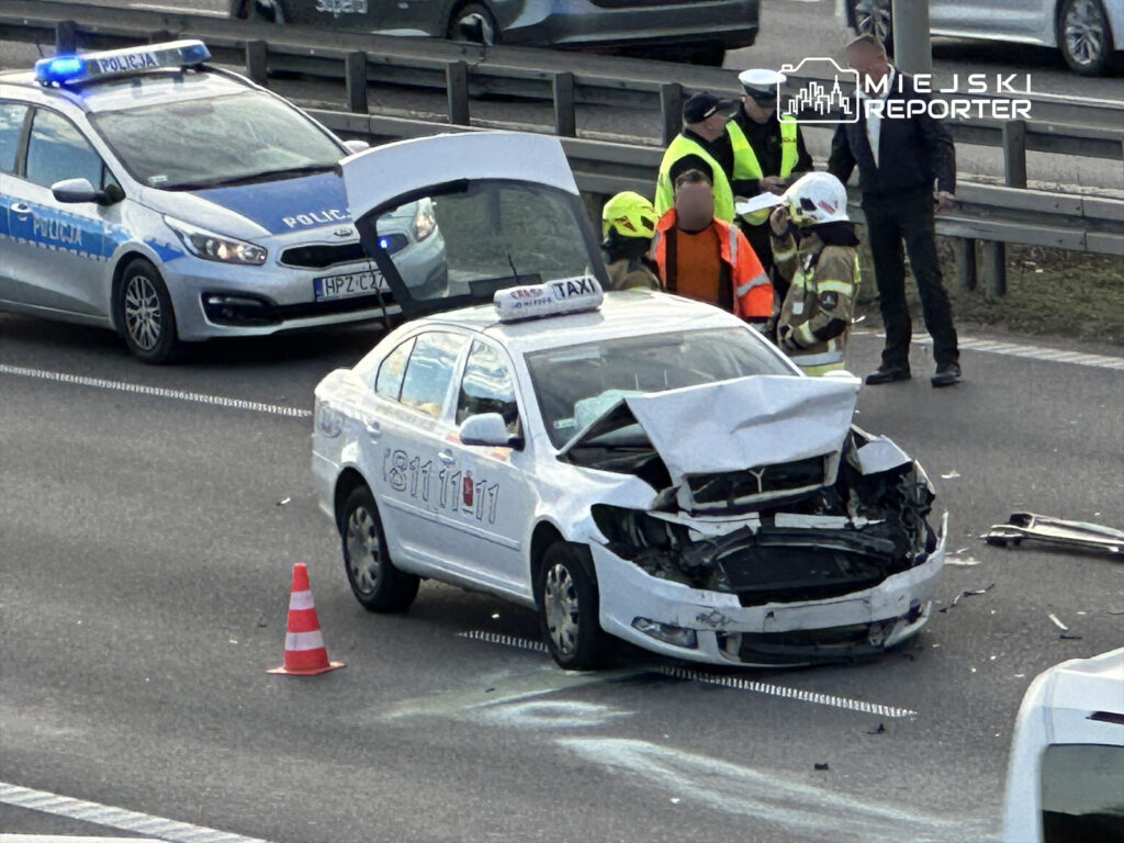 W miejscu wypadku drogowego stoi uszkodzony samochód taxi z wgniecionym przodem, obok pojazd policyjny z włączonymi sygnałami.
