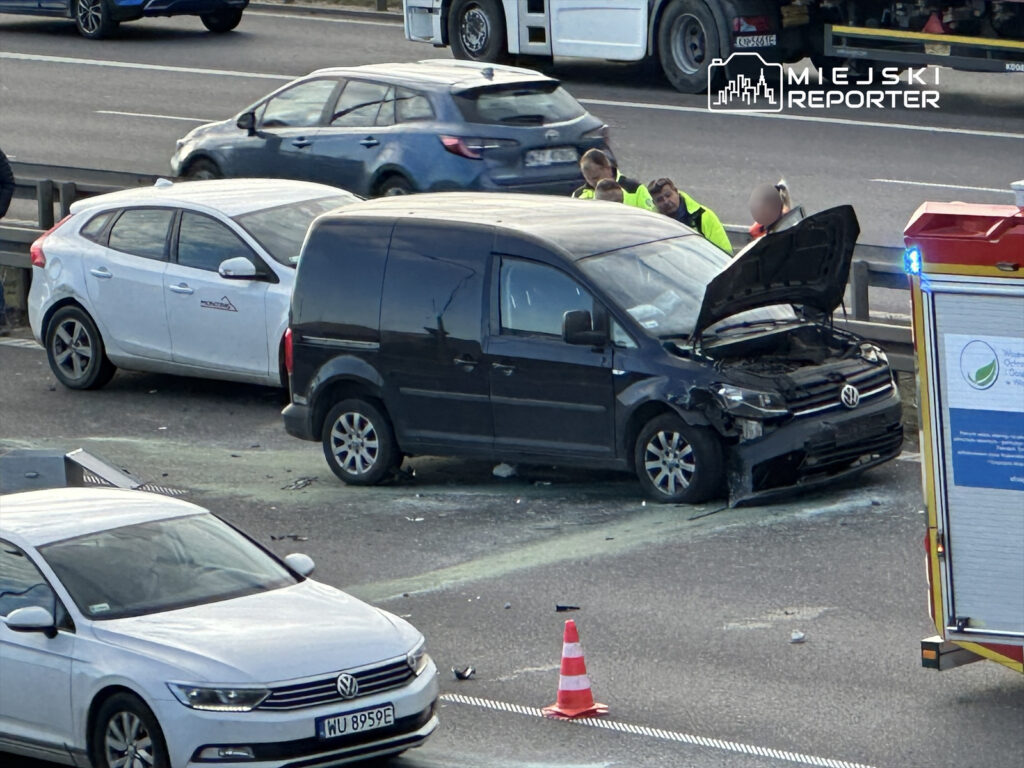 Czarny dostawczy samochód z otwartą maską stoi na jezdni po kolizji, obok dwóch innych pojazdów oraz ekipy ratunkowej.