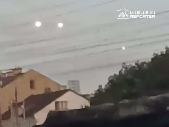 Na niebie widoczne są trzy jasne punkty świetlne, a w tle widać dachy budynków i linie energetyczne.