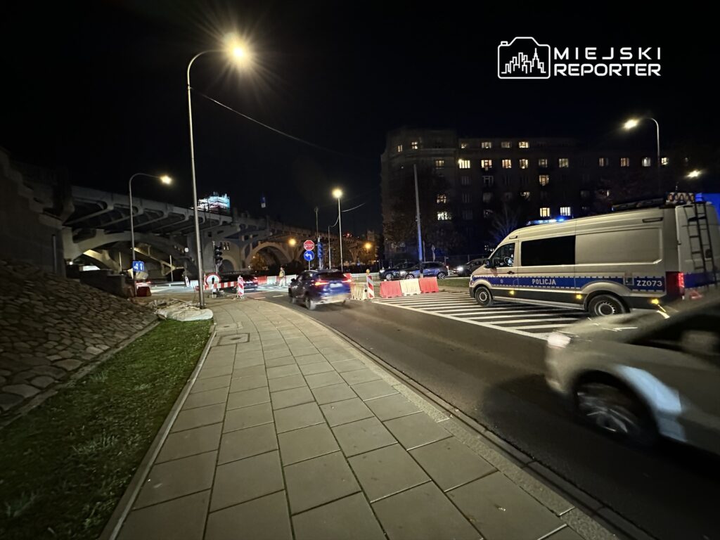 Policyjny radiowóz zaparkowany obok barier zabezpieczających na ruchliwej ulicy w nocy, w tle oświetlony most.