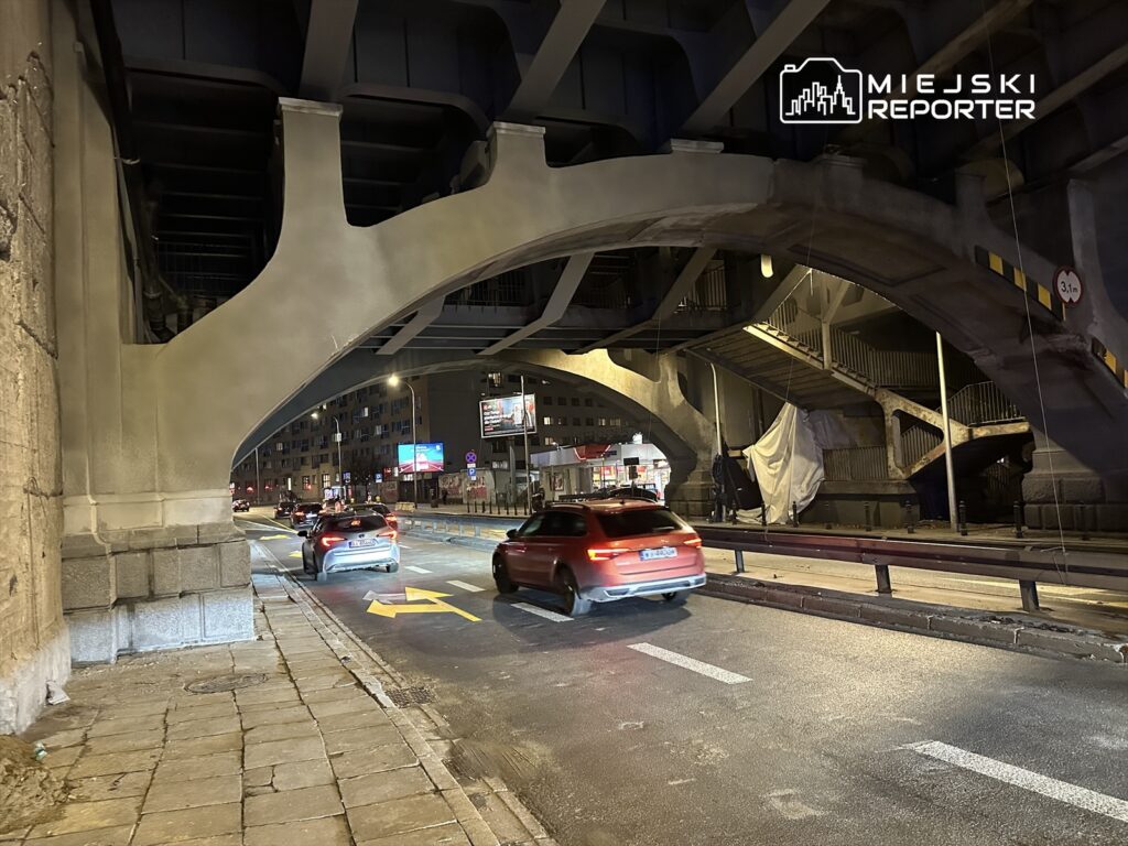 Ruch samochodowy pod mostem, z widocznymi schodami i reklamami w tle, w nocy w miejskim otoczeniu.