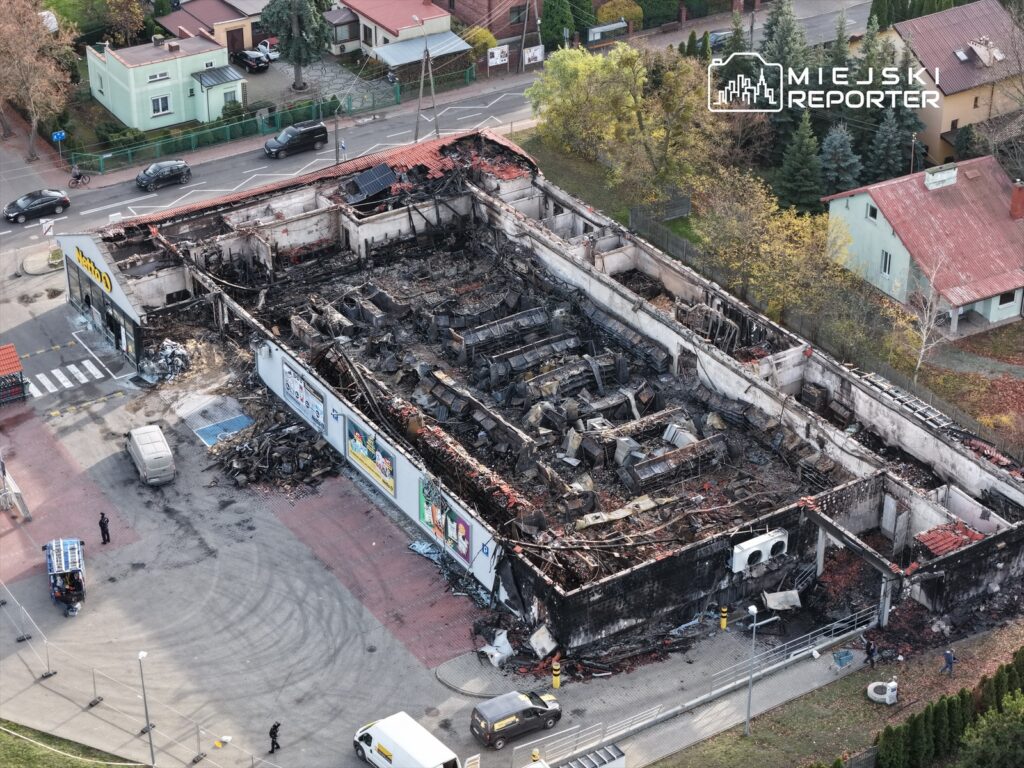 Zniszczony budynek handlowy z wypaloną konstrukcją, otoczony przez pojazdy i osoby w mundurach, w miejskim otoczeniu.