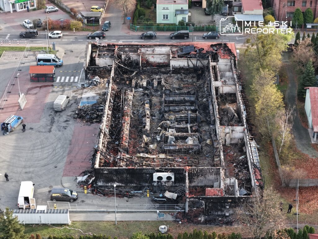 Zniszczony budynek po pożarze, z widocznymi resztkami konstrukcji i popiołem, otoczony przez pojazdy i osoby w mundurach.