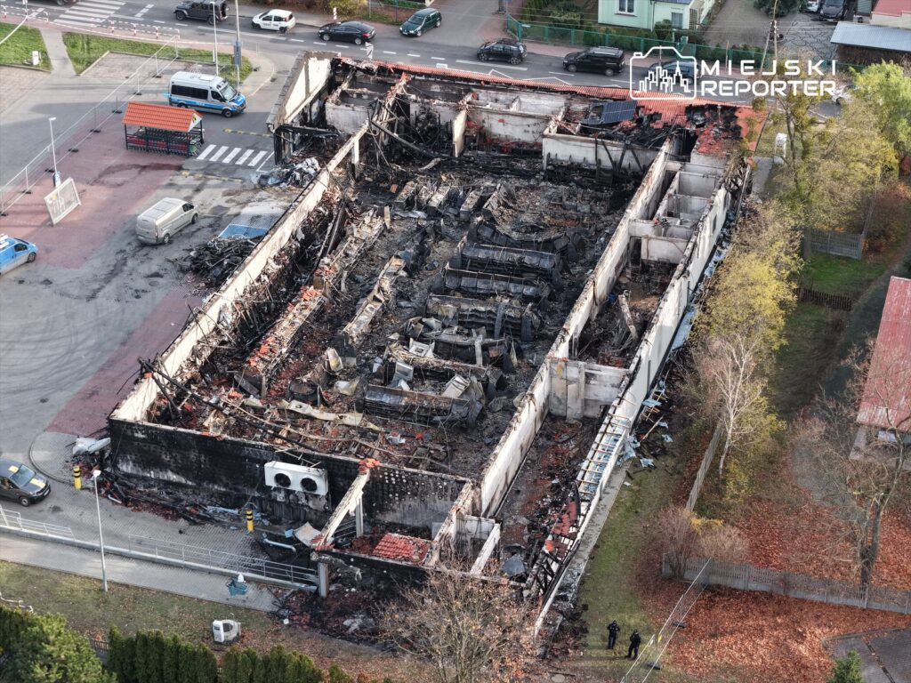 Zniszczony budynek z wypaloną strukturą dachową i ścianami, otoczony fragmentami gruzów oraz pojazdami na pobliskim parkingu.