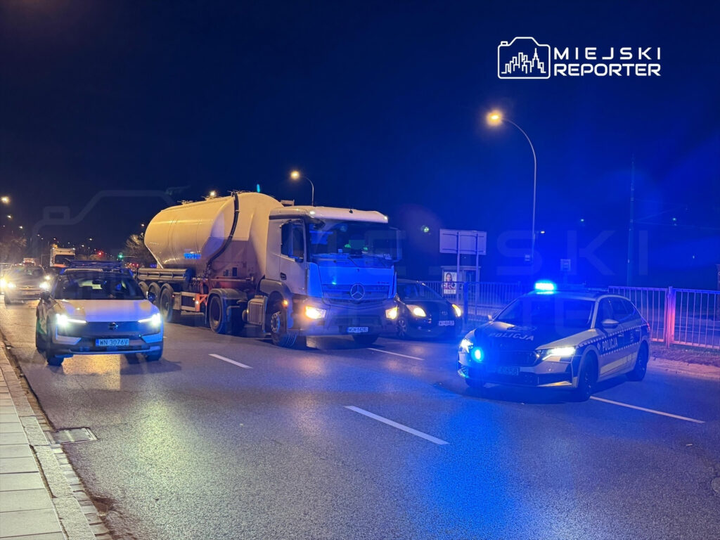Policyjny radiowóz z włączonymi sygnałami świetlnymi zatrzymany obok ciężarówki na ruchliwej ulicy nocą.