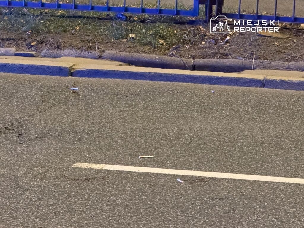 Na jezdni leżą papierosy obok krawężnika, w tle widoczny niebieski płot.