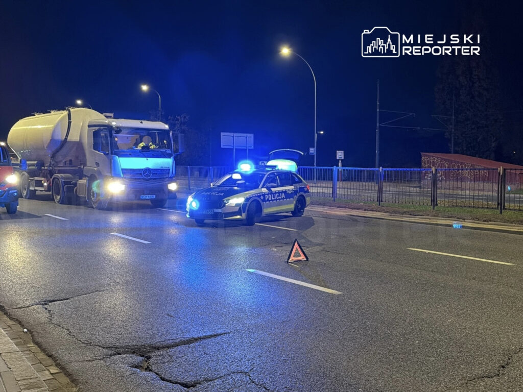 Policyjny radiowóz z włączonymi sygnałami świetlnymi blokuje drogę, obok stoi ciężarówka z cysterną.