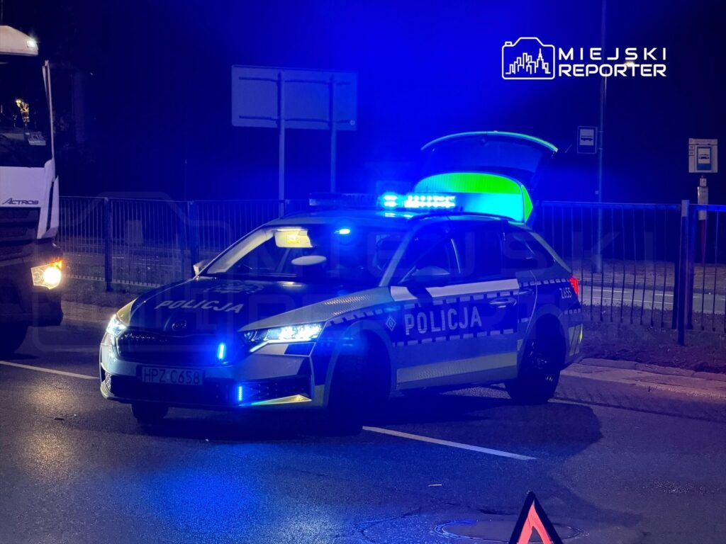 Policyjny radiowóz z włączonymi niebieskimi światłami zatrzymany na drodze nocą, obok ciężarówki.